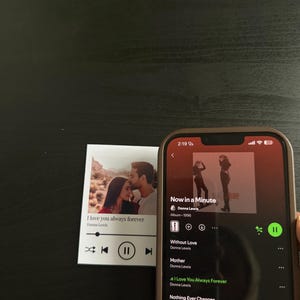 May include: A smartphone displays a music playlist with the song "Now in a Minute" by Donna Lewis. Beside it is a small print with the text "I love you always forever" and a photo of a couple.