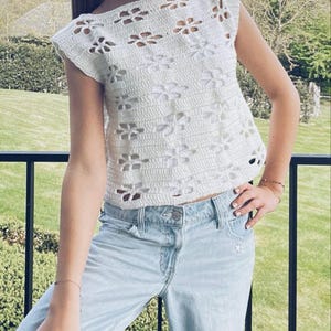May include: A white crochet top with a floral pattern and short sleeves. The top is worn with light blue jeans.