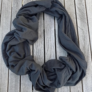 May include: A black, soft, and lightweight scarf made of a textured fabric. The scarf is folded and draped on a wooden surface.