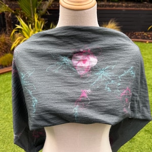 May include: A dark gray shawl with a floral design in pink and teal. The shawl is draped over a mannequin, showcasing the fabric's texture and the delicate pattern. The background features green grass and foliage.