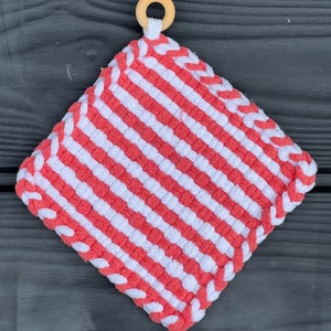 May include: A pink and white striped pot holder with a wooden loop handle. The pot holder is made of woven fabric and has a diamond shape.