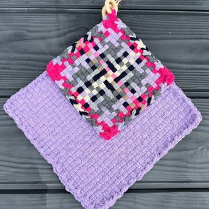 May include: A woven pot holder and matching square potholder. The pot holder features a woven design in pink, gray, black, and white, with a wooden ring for hanging. The potholder is a solid lavender color.