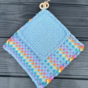 May include: Two blue knitted pot holders, one with a colorful checkered border and the other with a plain design. The pot holders are hanging from a wooden ring.