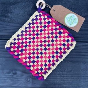 Puede incluir: Un posavasos cuadrado tejido con un patrón de cuadros rosa, morado y crema. El posavasos tiene un anillo de madera unido a la parte superior y una etiqueta marrón con el texto "Never Not Weaving" en blanco.