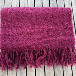 Puede incluir: Bufanda magenta doblada, con un aspecto texturizado y difuso, con flecos en el borde inferior. La bufanda está hecha de un material suave, adecuado para el frío. El color es rosa intenso.