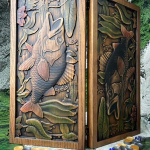 Puede incluir: Un juego de backgammon de madera con un relieve tallado de peces y plantas acuáticas. El juego está abierto, revelando la superficie de juego y las piezas. Los peces están detallados con aletas y escamas, y las plantas son verdes y marrones.