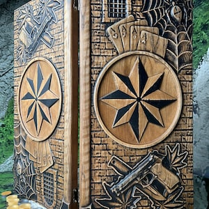 May include: A wooden game box with intricate carvings. The box features a compass rose design, playing cards, a spider web, and a gun. The box is open, revealing a set of checkers and dice. The box is made of wood and has a natural finish.