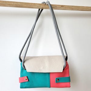 May include: A color-blocked crossbody bag with a cream-colored flap, teal and coral body, and gray rope strap. The bag features a rectangular shape with a snap closure on the front. The bag is hanging from a wooden rod.
