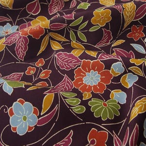 May include: A dark purple fabric featuring a repeating floral pattern with stylized flowers and leaves in shades of orange, light blue, yellow, and green. The textile has a smooth texture and is draped to show folds across the surface.