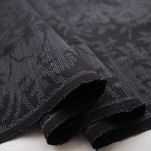 May include: A length of black fabric featuring a subtle, intricate floral pattern created with tiny white dots. The textile is folded over itself, revealing the woven texture and the consistent, repeating design across the dark surface.