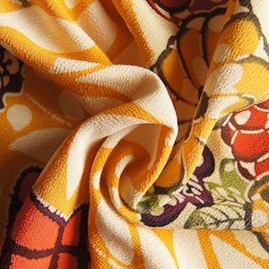 May include: Close-up of a folded fabric with a repeating floral pattern. The design features large, stylized flowers and butterfly-like shapes in shades of yellow, orange, red, purple, and cream. The fabric has a textured appearance.