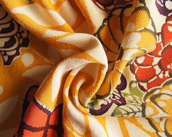 May include: Close-up of a folded fabric with a repeating floral pattern. The design features large, stylized flowers and butterfly-like shapes in shades of yellow, orange, red, purple, and cream. The fabric has a textured appearance.