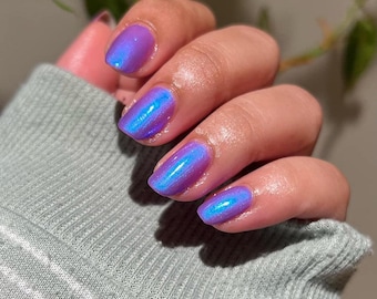 May include: Close-up of a hand with polished nails. The nails are painted with a purple iridescent polish that shifts to blue in the light. The hand is resting on a light green sweater.
