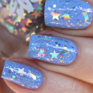 May include: Close-up of fingernails painted with light blue nail varnish. The varnish is decorated with iridescent star and crescent moon-shaped glitter. The nails have a glossy finish and are well-manicured. The overall effect is whimsical and celestial.