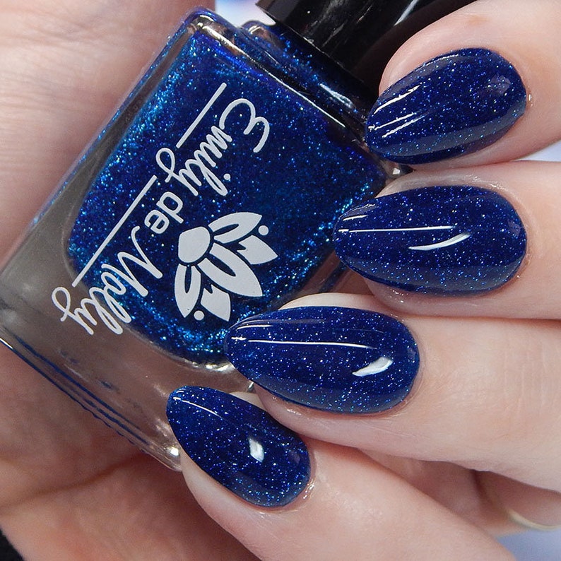 Pode incluir: Um frasco de vidro de verniz de unhas azul profundo com um acabamento de purpurina fina, apresentando o texto branco Emily de Molly e um log&oacute;tipo de flor de l&oacute;tus. O verniz &eacute; aplicado em unhas ovais, exibindo uma cor azul vibrante e brilhante.