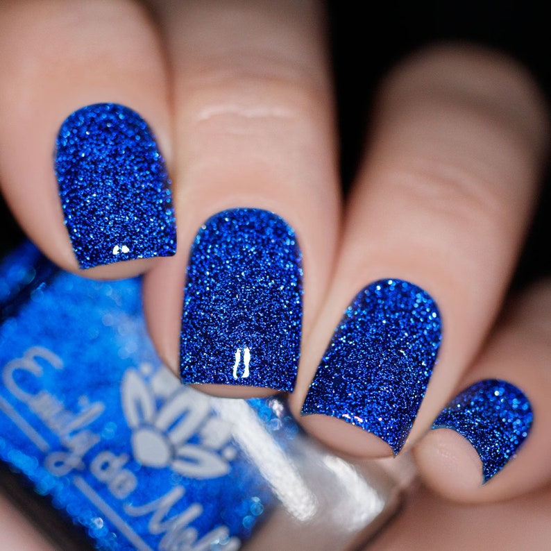 Pode incluir: Unhas pintadas com um verniz de purpurina azul profundo e vibrante. O verniz tem uma textura densa e brilhante que cobre toda a superf&iacute;cie da unha. Um frasco de vidro do mesmo verniz azul com purpurina e a marca Emily de Molly &eacute; vis&iacute;vel no canto inferior esquerdo.