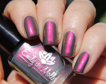 May include: A bottle of dark green nail polish with a silver label that says "Emily de Molly". The bottle is sitting in front of a hand with three fingernails painted with a dark green and pink iridescent nail polish.