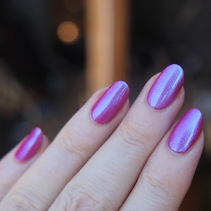 K&ouml;nnte beinhalten: Nahaufnahme einer Hand mit vier Fingern&auml;geln, die mit einem schimmernden rosa Nagellack mit einem blauen irisierenden Schimmer lackiert sind.