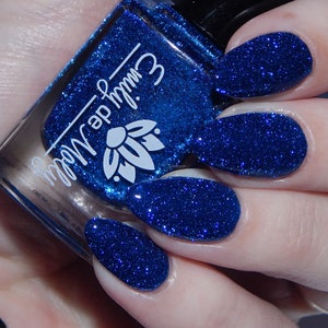 Pode incluir: Um frasco de vidro de verniz de unhas azul profundo com um acabamento de glitter denso, apresentando o texto branco "emily de molly" e um log&oacute;tipo de flor de l&oacute;tus na frente. O verniz &eacute; aplicado em unhas ovais, exibindo uma cor azul vibrante.
