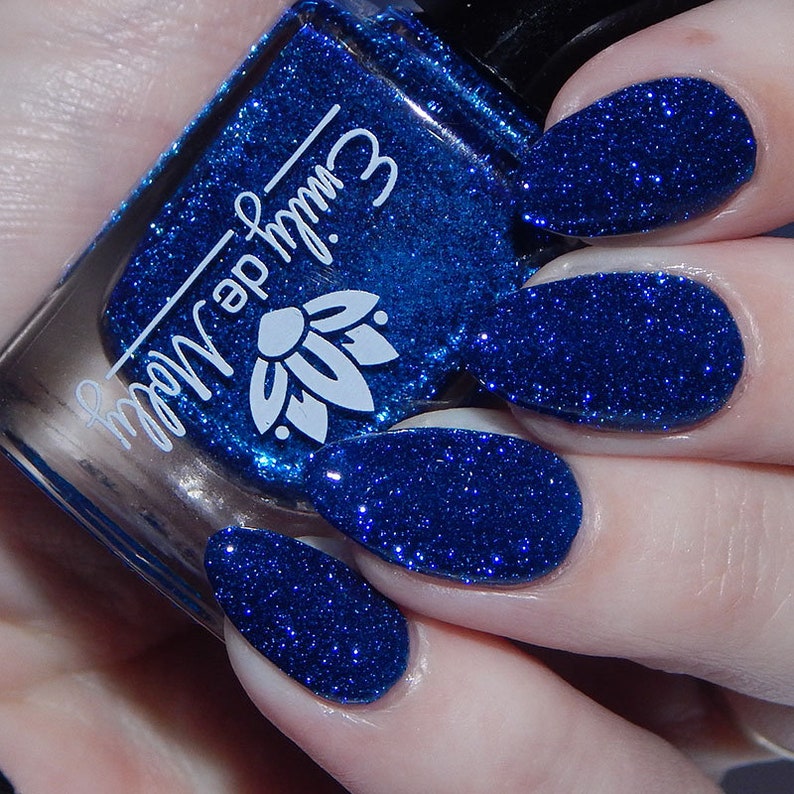 Pode incluir: Um frasco de vidro de verniz de unhas azul profundo com um acabamento de glitter denso, apresentando o texto branco "emily de molly" e um log&oacute;tipo de flor de l&oacute;tus na frente. O verniz &eacute; aplicado em unhas ovais, exibindo uma cor azul vibrante.