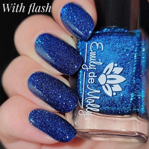 Pode incluir: Um frasco de vidro de verniz de unhas azul profundo com um acabamento brilhante e refletor. O r&oacute;tulo apresenta o texto branco 'Emily de Molly' e um &iacute;cone de flor de l&oacute;tus. O verniz est&aacute; aplicado nas unhas, exibindo uma textura azul vibrante e cintilante.