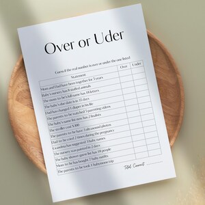 May include: A white paper with the text "Over or Uder" and a game for a baby shower. The game sheet is on a wooden plate. The game includes statements about the parents and baby.