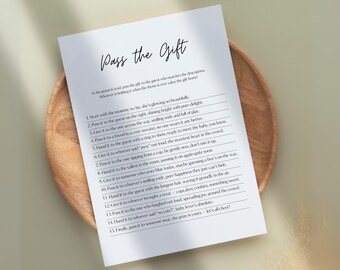 May include: A white paper with the text "Pass the Gift" in elegant script, listing instructions for a baby shower game. The paper rests on a round, light-colored wooden tray, with a neutral background.