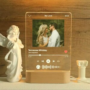 May include: A clear acrylic LED night light with a wooden base. The light displays a photo and the text "Tennessee Whiskey" by Chris Stapleton, resembling a music player interface. The light is plugged in and illuminated.