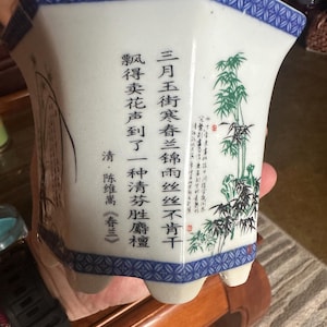 May include: A white ceramic planter with a blue patterned rim and base. The planter features Chinese calligraphy and a bamboo illustration. The planter has a unique shape and is held by a person.