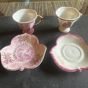 May include: Two antique teacups and saucers. One cup is pink with a gold handle, and the other is white with a floral design. The saucers are leaf-shaped, one pink and one white, both with gold accents. A vintage tea set.