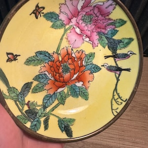 May include: A small, round, yellow dish with a gold rim. The dish is decorated with a floral design featuring pink and orange flowers, green leaves, and small birds. Two butterflies are also depicted on the dish.