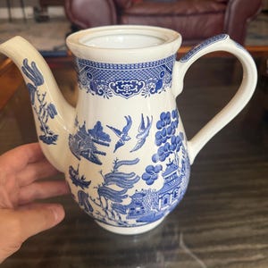 May include: A cream-colored ceramic teapot featuring a blue willow pattern. The design includes intricate illustrations of pagodas, willow trees, and birds in flight. The teapot has a curved handle, a tapered spout, and a wide opening at the top.