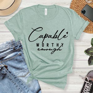 May include: A light green t-shirt with the words "Capable, Worthy, Enough" in black script. The shirt is made of soft material and is knotted at the bottom. A pair of blue jeans is visible on the left side of the shirt.