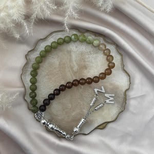May include: A prayer bead strand featuring a gradient of smooth spherical beads transitioning from olive green to dark brown. The strand includes a silver-toned metal tassel component with intricate detailing and two silver letter charms, Z and M.