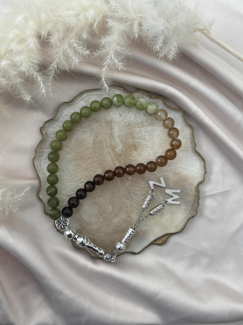 May include: A prayer bead strand featuring a gradient of smooth spherical beads transitioning from olive green to dark brown. The strand includes a silver-toned metal tassel component with intricate detailing and two silver letter charms, Z and M.
