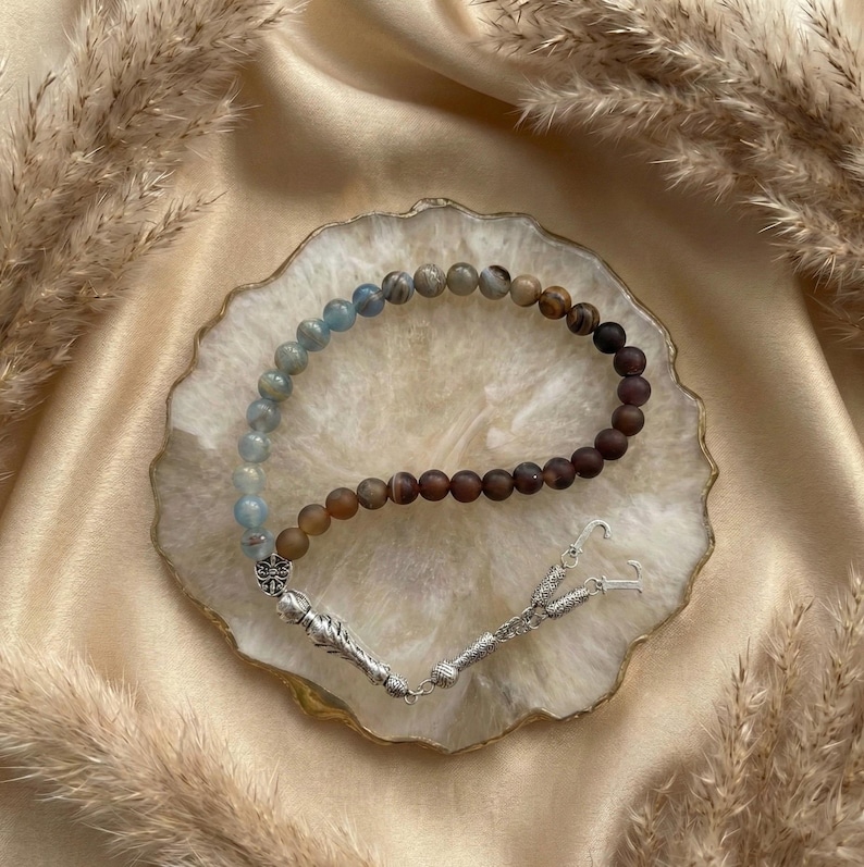 May include: A prayer bead strand featuring a gradient of spherical beads transitioning from light blue to dark brown. The strand includes a silver-toned metal decorative spacer and a silver-toned metal tassel component with two hook-shaped ends.