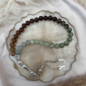 May include: A prayer bead strand featuring a gradient of polished spherical beads transitioning from dark brown to light green. The strand includes a silver-toned metal ornament with a fleur-de-lis design, a textured silver tassel piece, and two silver letter charms, J and Y.