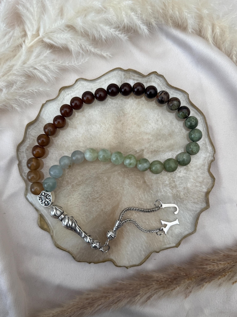 May include: A prayer bead strand featuring a gradient of polished spherical beads transitioning from dark brown to light green. The strand includes a silver-toned metal ornament with a fleur-de-lis design, a textured silver tassel piece, and two silver letter charms, J and Y.