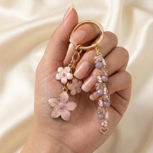 May include: A gold-colored keychain with a cluster of pink flower charms and a beaded tassel. The tassel features lavender and white beads, and a small heart charm. The keychain is held in a hand.