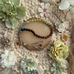 May include: A prayer bead strand featuring a gradient of smooth, round beads transitioning from light green to dark brown. The strand includes silver-toned metal spacers and a silver-toned tassel component with a dangling letter B charm.