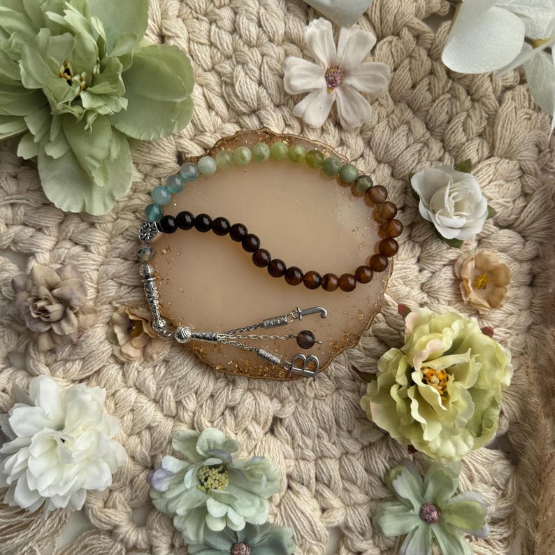 May include: A prayer bead strand featuring a gradient of smooth, round beads transitioning from light green to dark brown. The strand includes silver-toned metal spacers and a silver-toned tassel component with a dangling letter B charm.