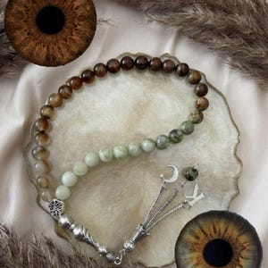 May include: A prayer bead strand featuring a gradient of smooth, round beads transitioning from dark brown to light green. The strand includes a silver-toned metal tassel with a crescent moon charm and a letter K charm, finished with a decorative metal bead.