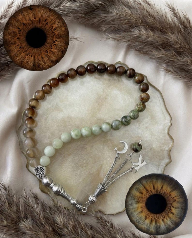 May include: A prayer bead strand featuring a gradient of smooth, round beads transitioning from dark brown to light green. The strand includes a silver-toned metal tassel with a crescent moon charm and a letter K charm, finished with a decorative metal bead.