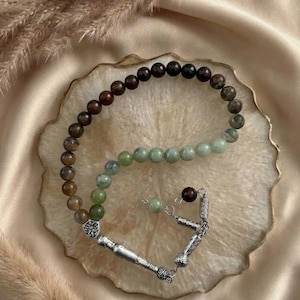 May include: A prayer bead strand featuring a gradient of smooth, round beads transitioning from dark brown to light green. The strand is finished with a silver-toned metal tassel component that includes a decorative floral-shaped spacer bead.