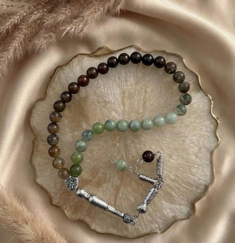 May include: A prayer bead strand featuring a gradient of smooth, round beads transitioning from dark brown to light green. The strand is finished with a silver-toned metal tassel component that includes a decorative floral-shaped spacer bead.