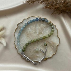 May include: A prayer bead strand featuring a gradient of smooth, round beads transitioning from light blue to olive green. The strand is finished with ornate silver-toned metal components, including a decorative shield-shaped charm and tassels.