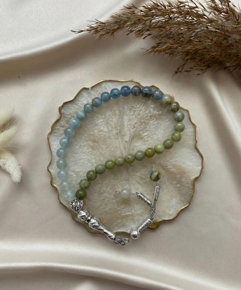 May include: A prayer bead strand featuring a gradient of smooth, round beads transitioning from light blue to olive green. The strand is finished with ornate silver-toned metal components, including a decorative shield-shaped charm and tassels.