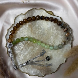 May include: A prayer bead strand featuring a mix of smooth brown, green, and marbled stone beads. The strand includes a silver-toned metal ornament with a fleur-de-lis design, a feather-shaped charm, and two silver-toned letter charms, E and T.
