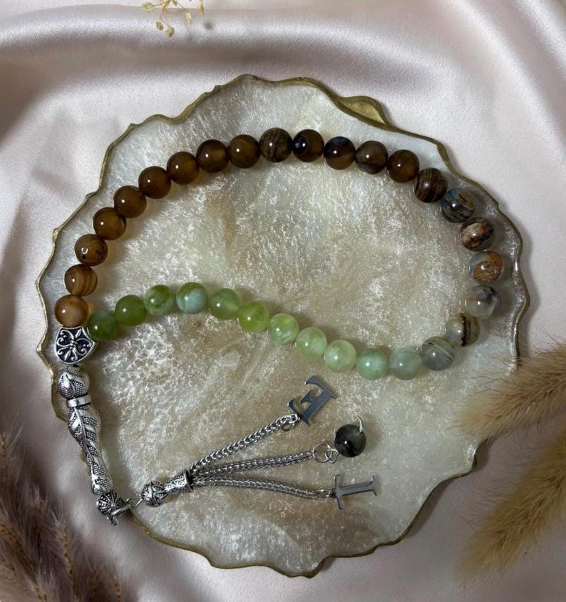 May include: A prayer bead strand featuring a mix of smooth brown, green, and marbled stone beads. The strand includes a silver-toned metal ornament with a fleur-de-lis design, a feather-shaped charm, and two silver-toned letter charms, E and T.