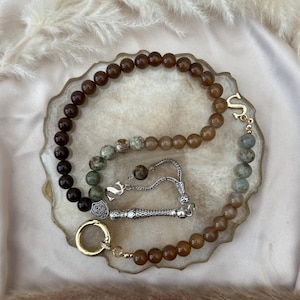 May include: A prayer bead strand featuring a mix of dark brown, tan, and mottled green stone beads. The strand includes a silver-toned metal tassel, a silver-toned letter S charm, and a gold-toned circular ring clasp at the end of the bead loop.